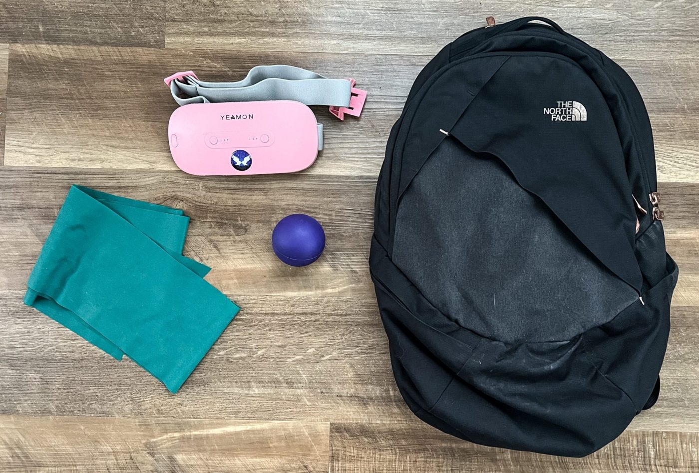 Black backpack, purple lacrosse ball, green thera band, and pink portable heating pad laid flat on a wood floor.