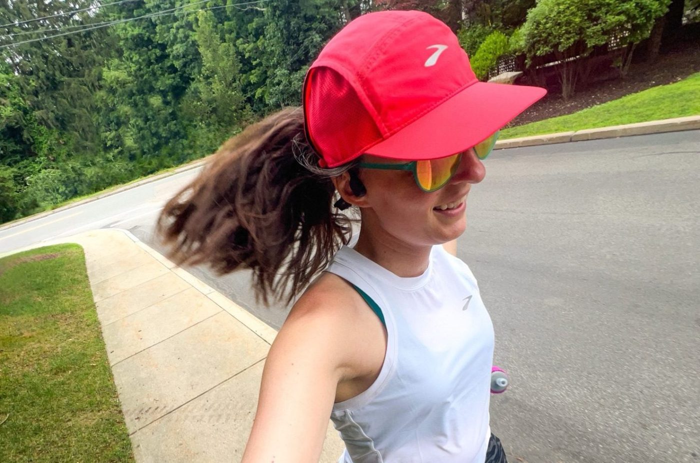 Woman running in a neon pink Brooks Running hat and white tank top.